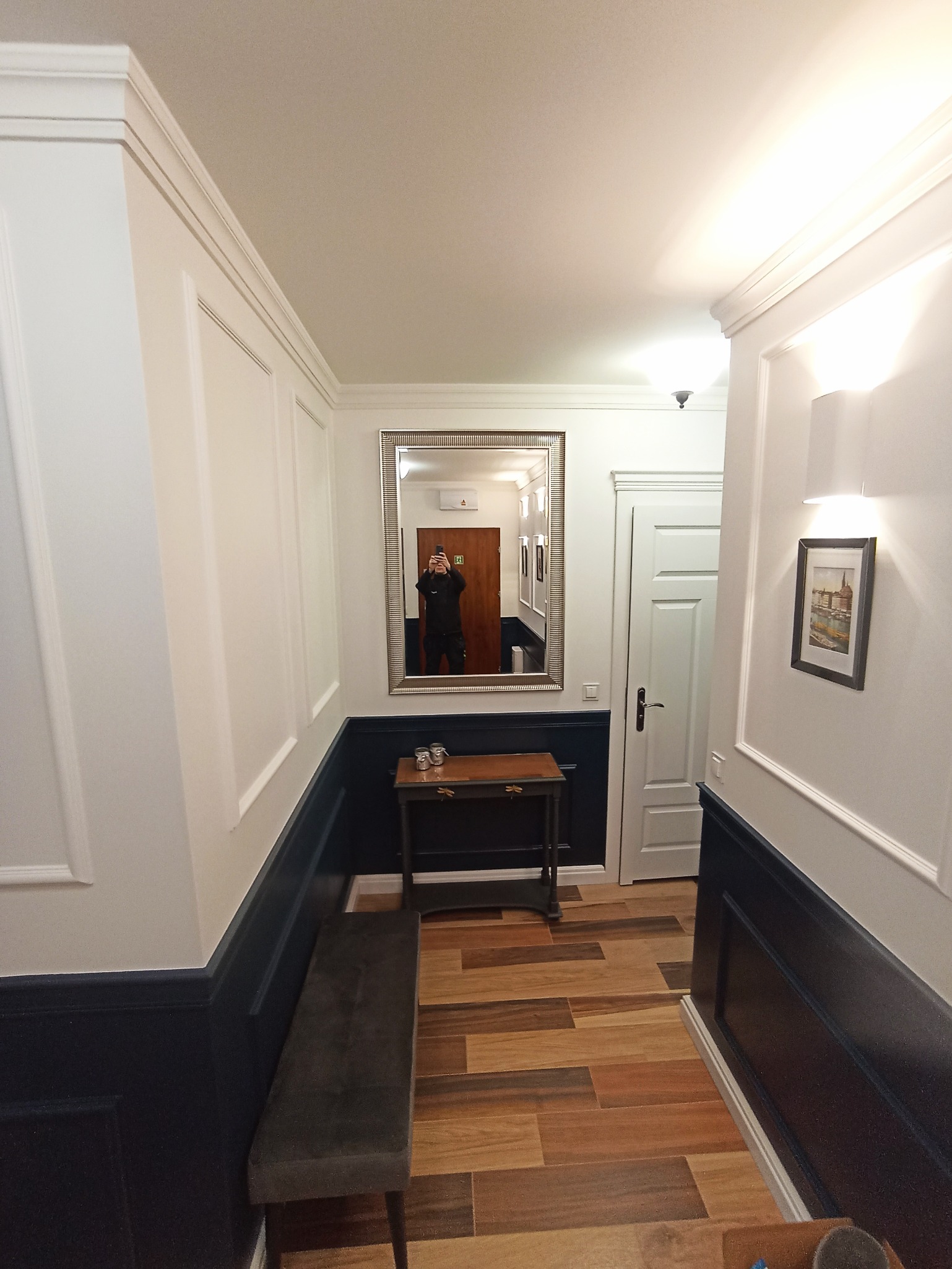 Korytarz po remoncie: białe ściany z ciemnoniebieską lamperią, drewnopodobne płytki, lustro w ozdobnej ramie i mała konsola. Widoczne detale wykończenia.