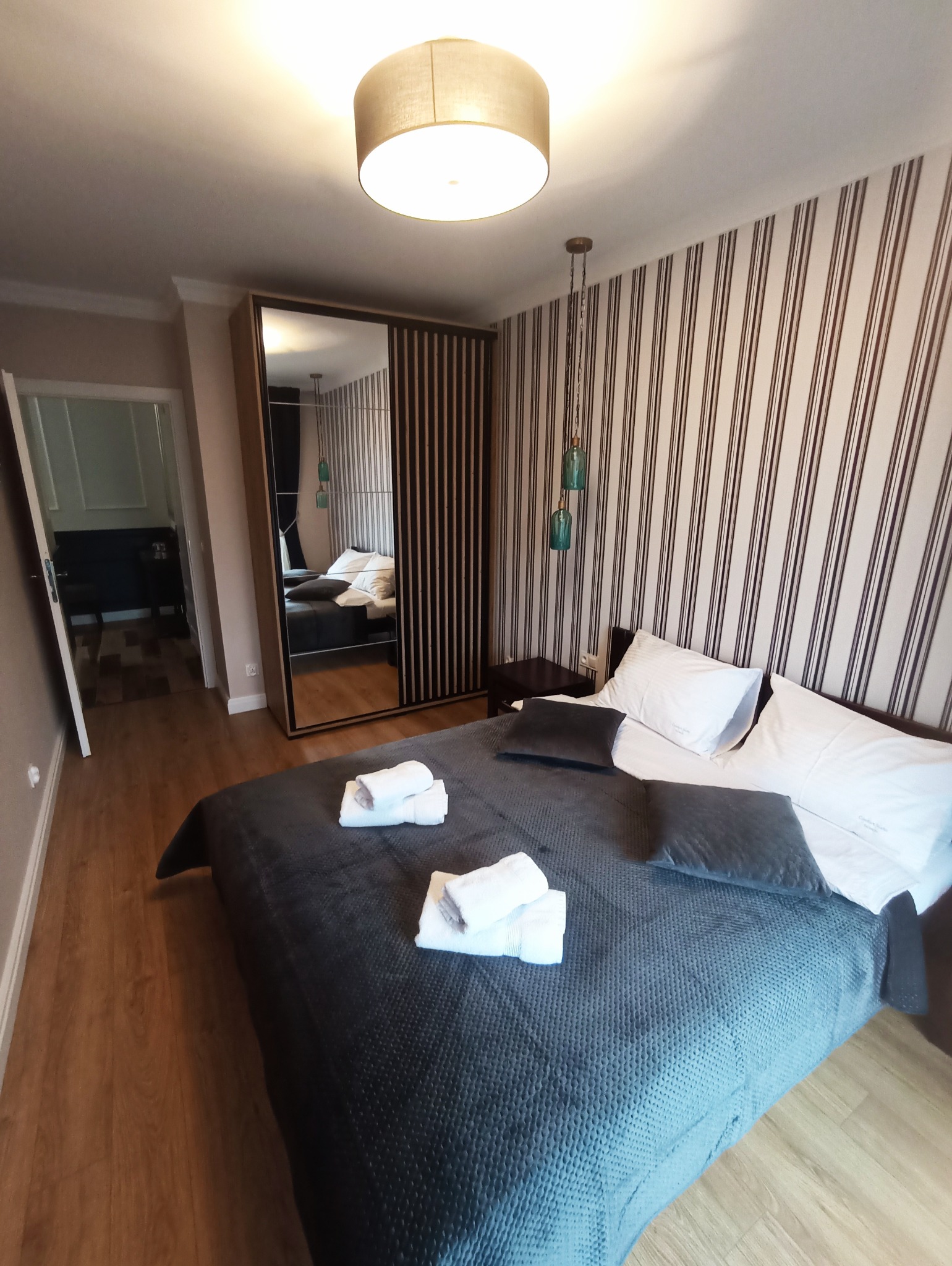 Nowoczesna sypialnia z łóżkiem, szafą z lustrem i tapetą w paski. Dwa dekoracyjne, szklane wisiorki zwisają z sufitu, dodając wnętrzu unikalnego charakteru.