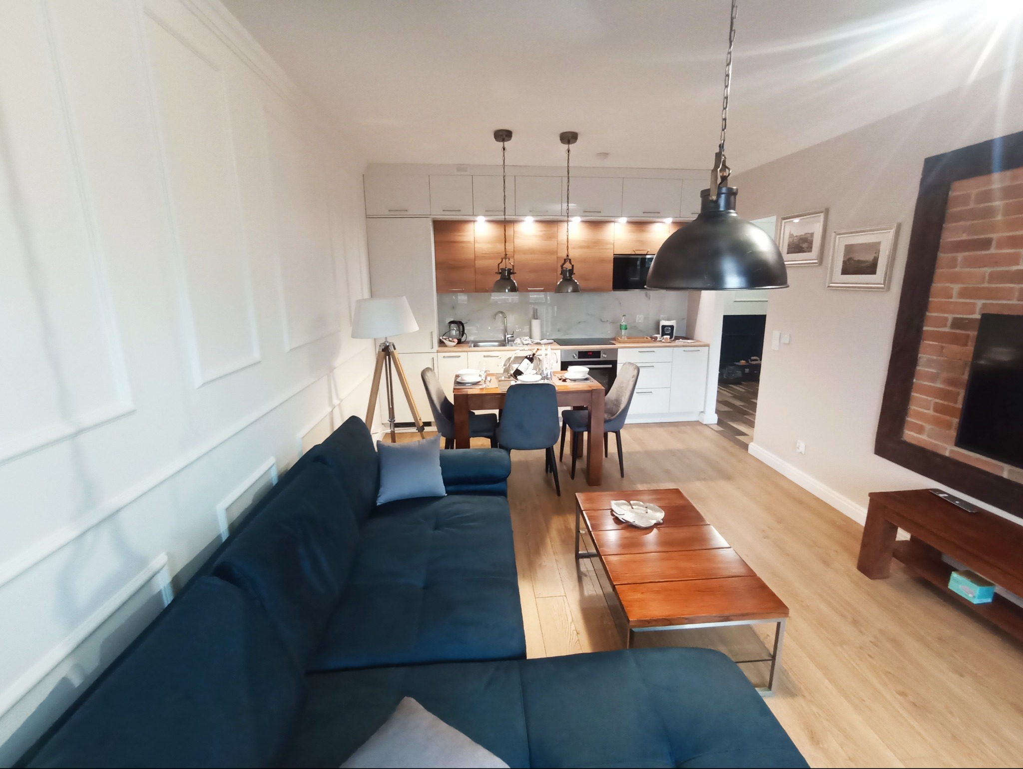 Przestronne wnętrze: salon z aneksem kuchennym, ciemna sofa, drewniany stolik kawowy, stół jadalny, białe ściany z dekoracyjnymi panelami i ceglana ściana z telewizorem.