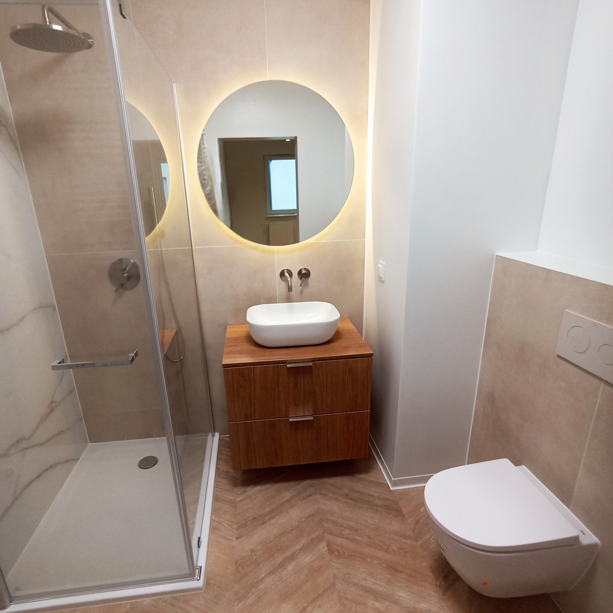 Nowoczesna łazienka po remoncie: kabina prysznicowa ze srebrnym panelem, umywalka na szafce z drewnianym fornirem, okrągłe lustro z podświetleniem LED i podwieszana toaleta.