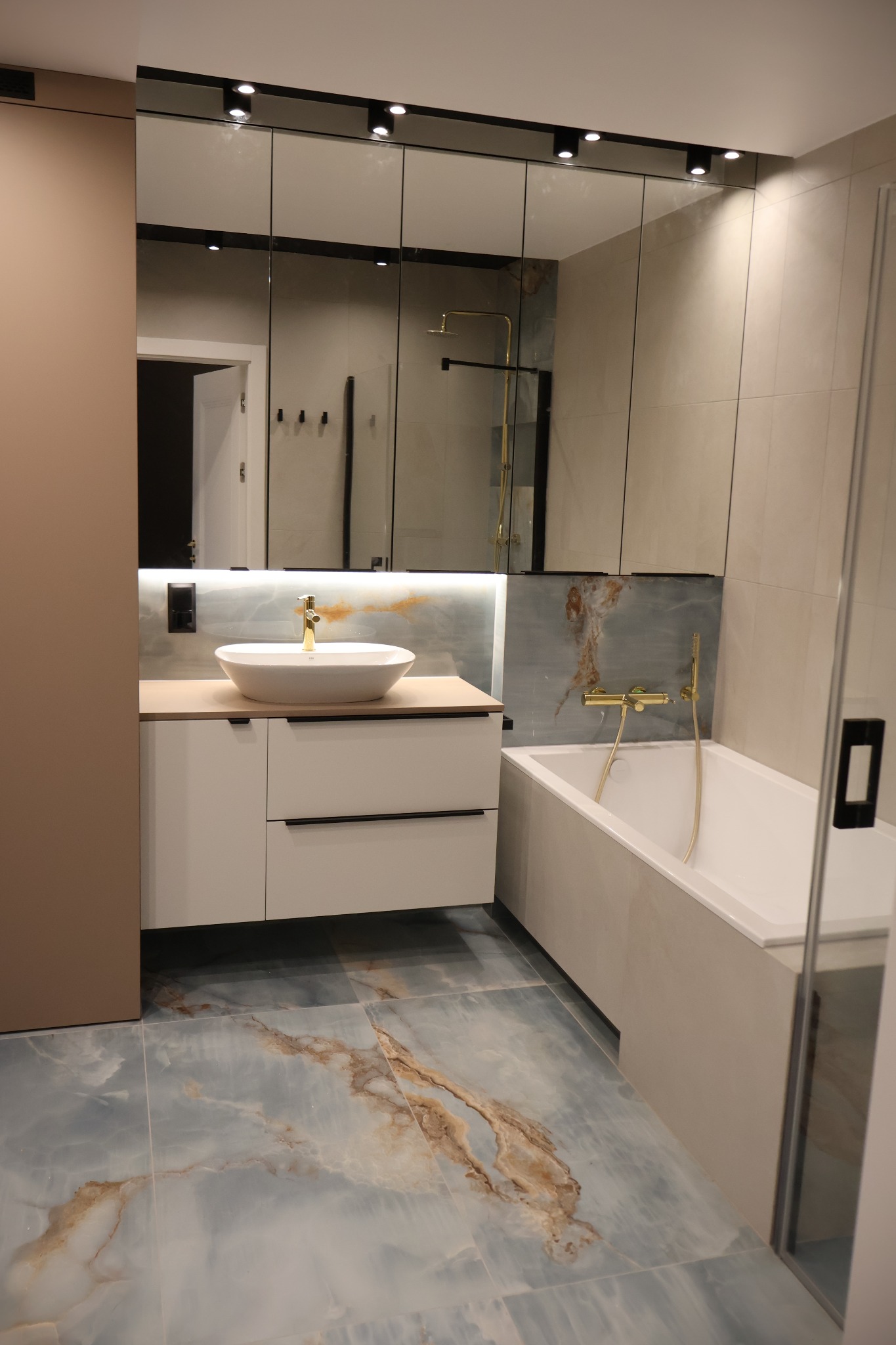 Nowoczesna łazienka z umywalką nablatową, lustrem z oświetleniem punktowym i wanną. Płytki podłogowe imitujące marmur w odcieniach błękitu i beżu.