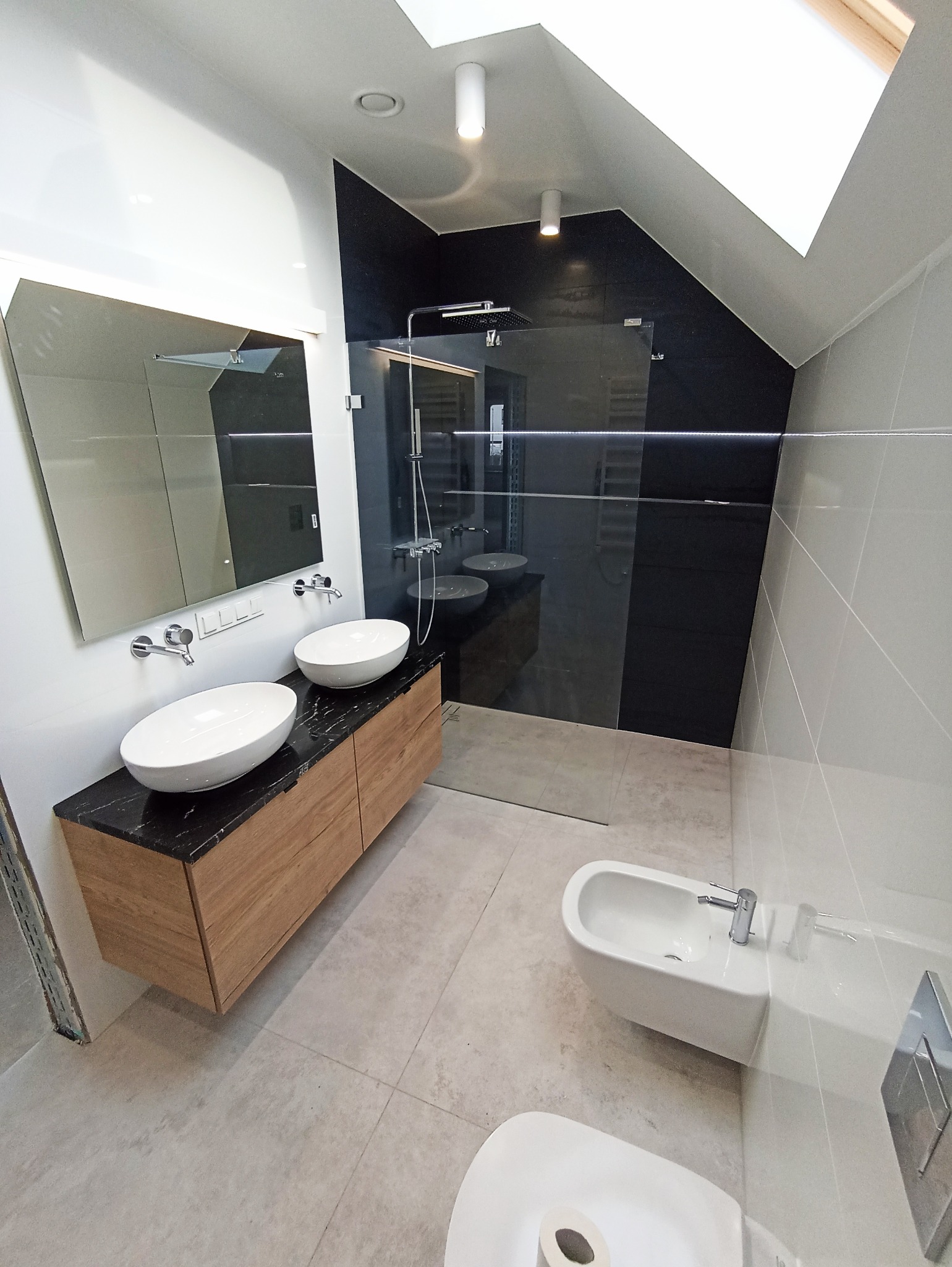 Nowoczesna łazienka na poddaszu z kabiną prysznicową typu walk-in, umywalkami nablatowymi na drewnianej szafce z czarnym blatem i oknem dachowym.