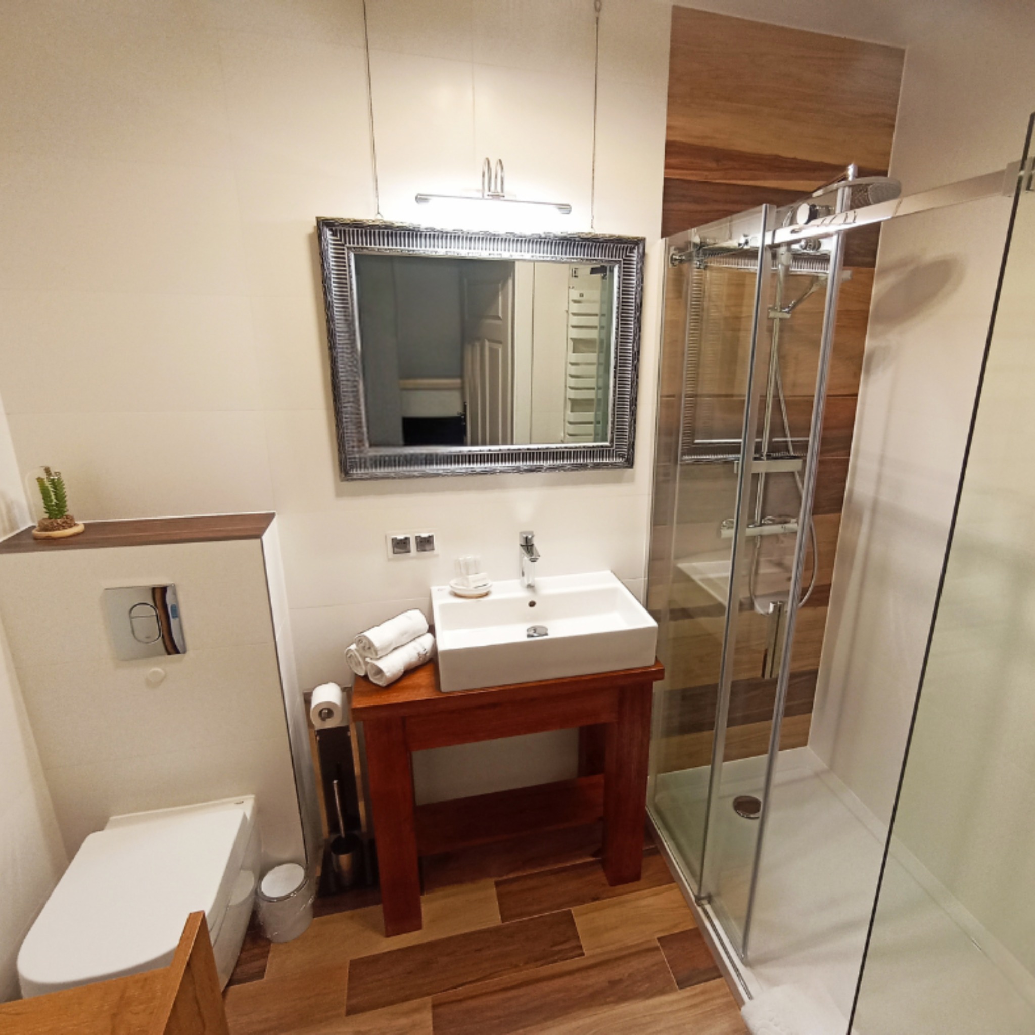 Nowoczesna łazienka z drewnianymi akcentami: kabina prysznicowa, umywalka na drewnianym postumencie, lustro w ozdobnej ramie i drewnopodobna podłoga.