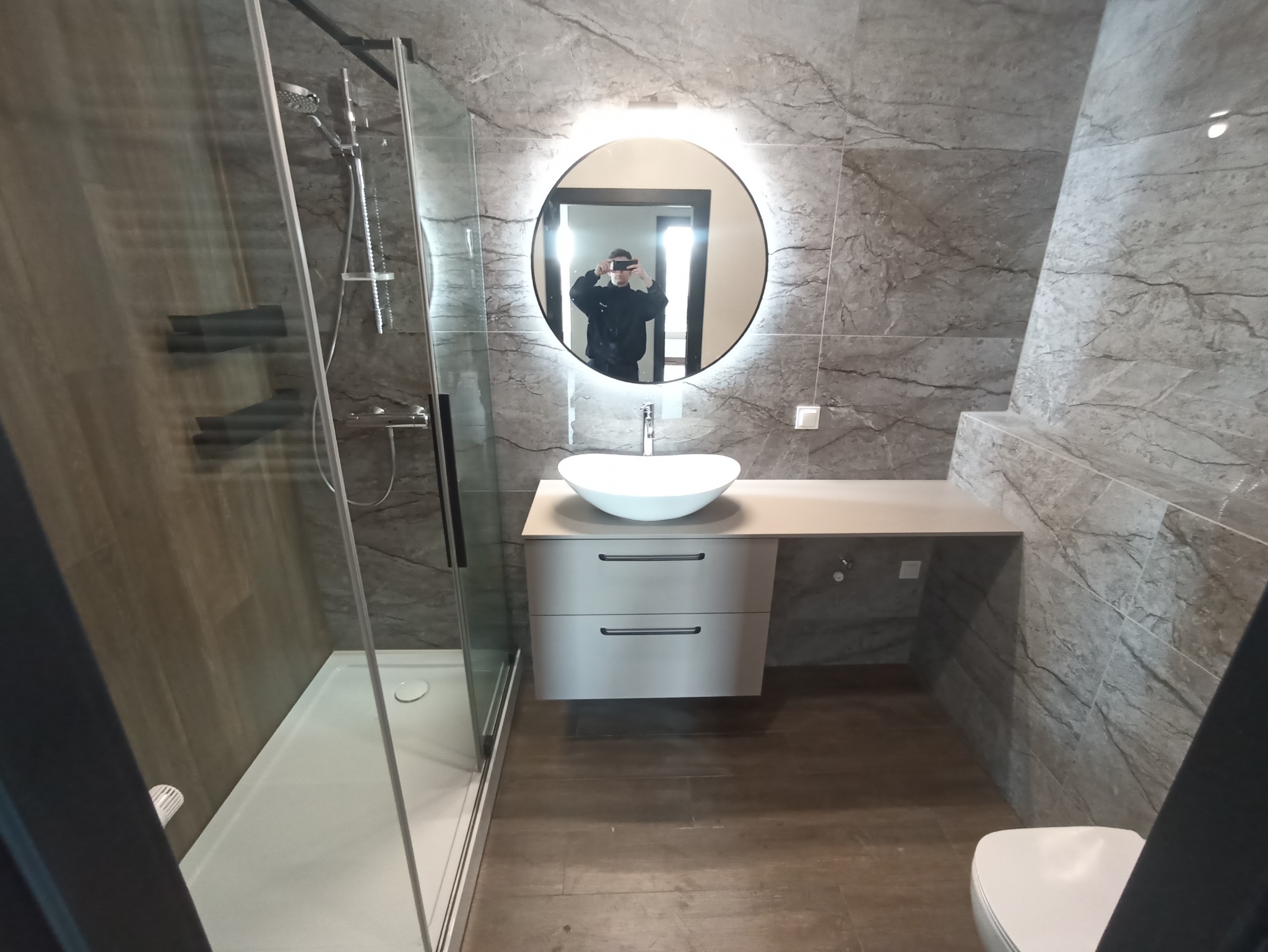 Nowoczesna łazienka w szarościach: kabina prysznicowa ze szklanymi drzwiami, umywalka na szafce z szufladami, okrągłe lustro z podświetleniem LED i marmurowe płytki.
