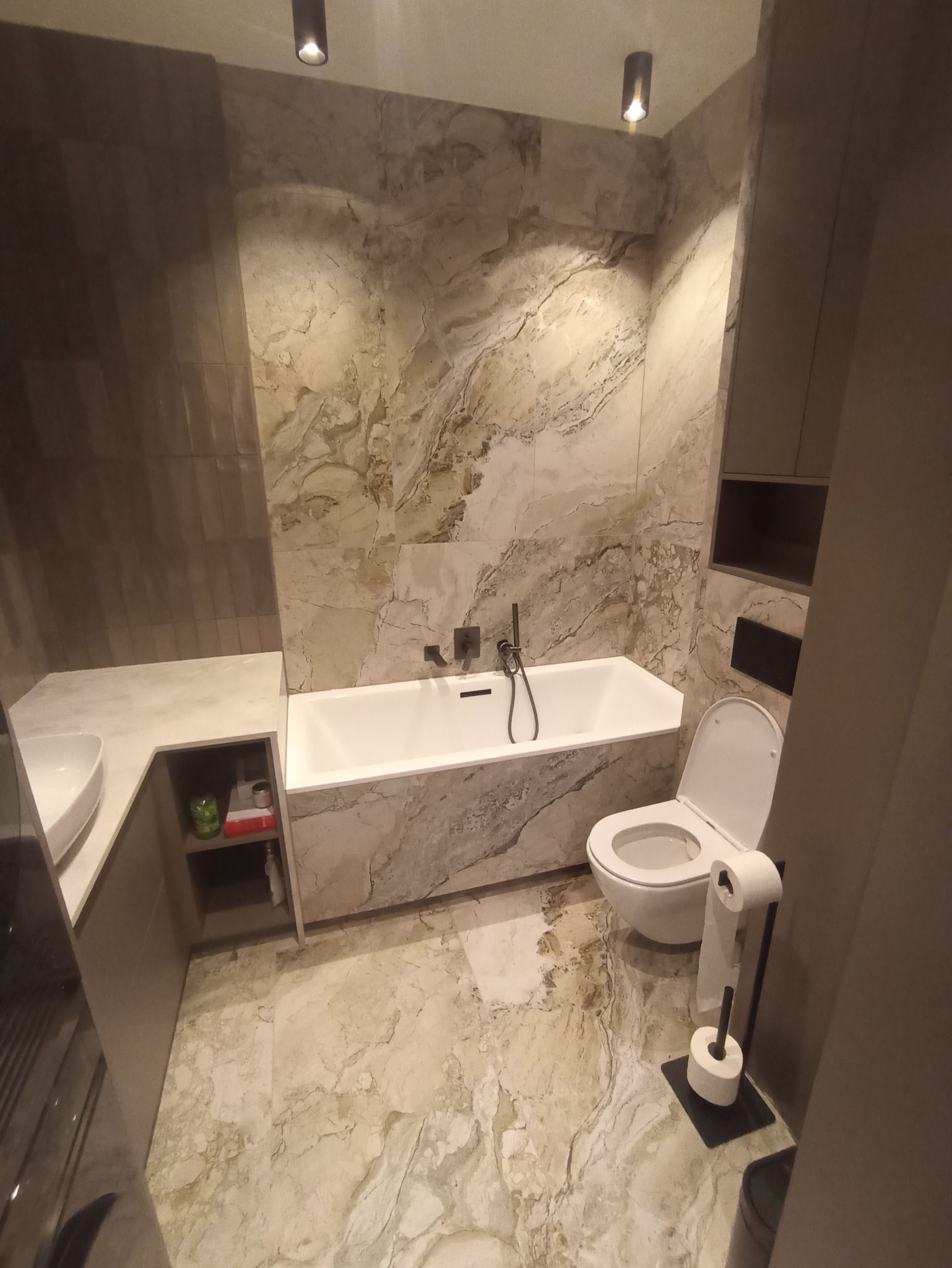 Elegancka łazienka z wanną, umywalką i toaletą, wykończona płytkami imitującymi marmur. Nowoczesne oświetlenie punktowe i minimalistyczne meble w odcieniach szarości.