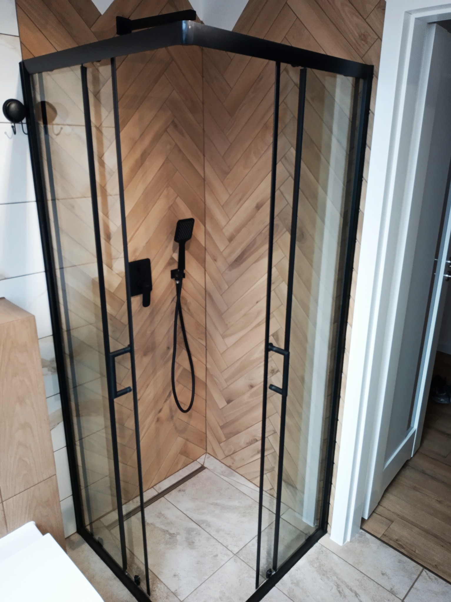 Kabina prysznicowa z czarnymi profilami na tle ściany wyłożonej drewnopodobną płytką w jodełkę. Widoczna słuchawka prysznicowa i jasna podłoga.