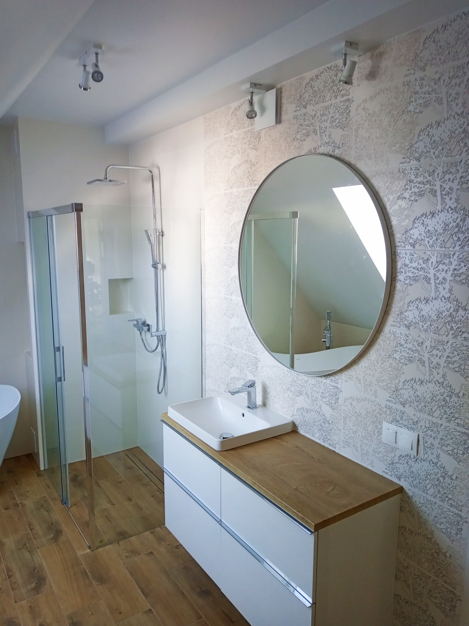 Nowoczesna łazienka: kabina prysznicowa ze szklanymi drzwiami, szafka z umywalką, okrągłe lustro i płytki z motywem drzew. Drewnopodobna podłoga.