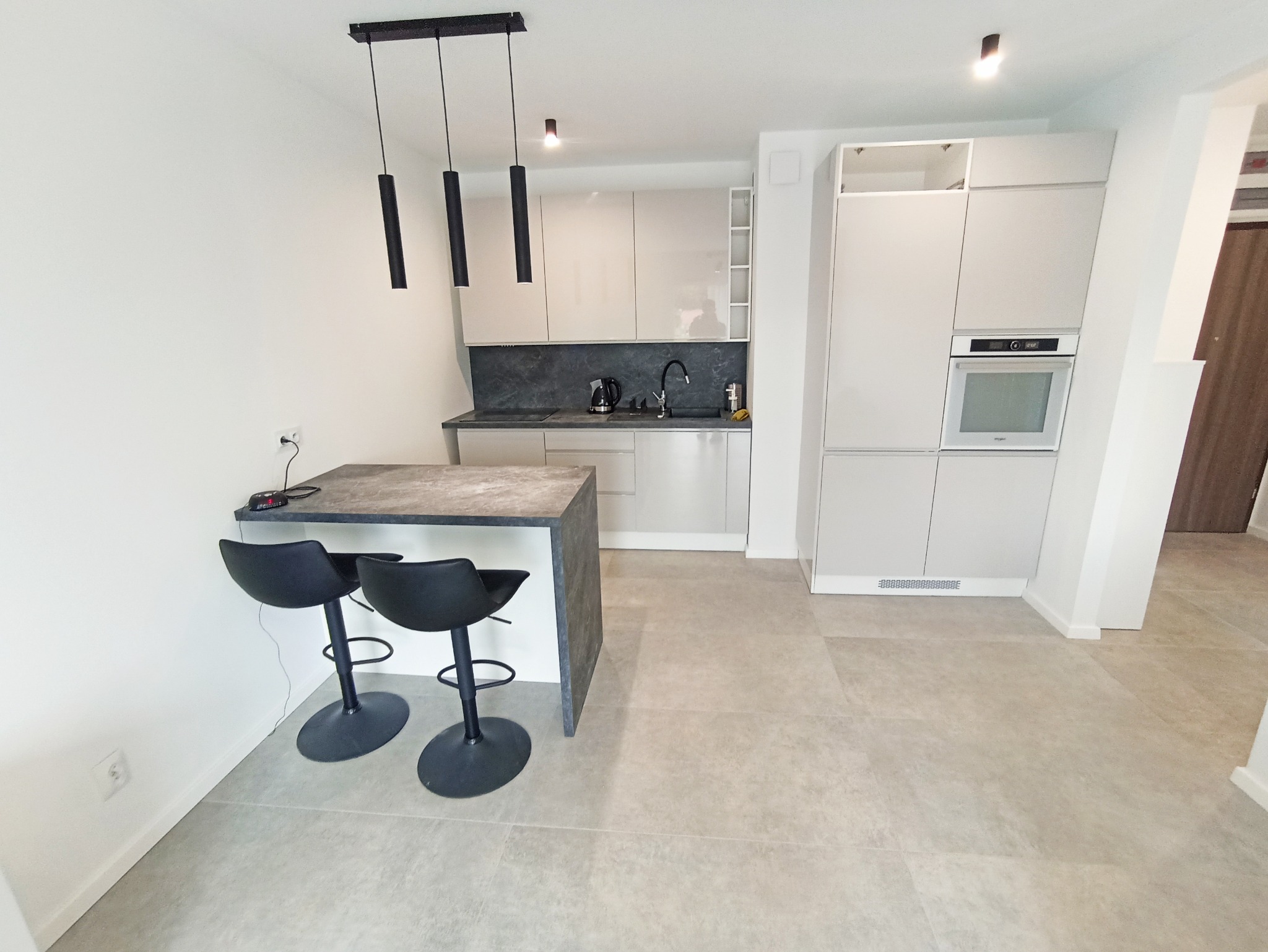 Jasna kuchnia z wyspą i dwoma czarnymi hokerami. Minimalistyczny design, szare płytki podłogowe, nowoczesne oświetlenie.