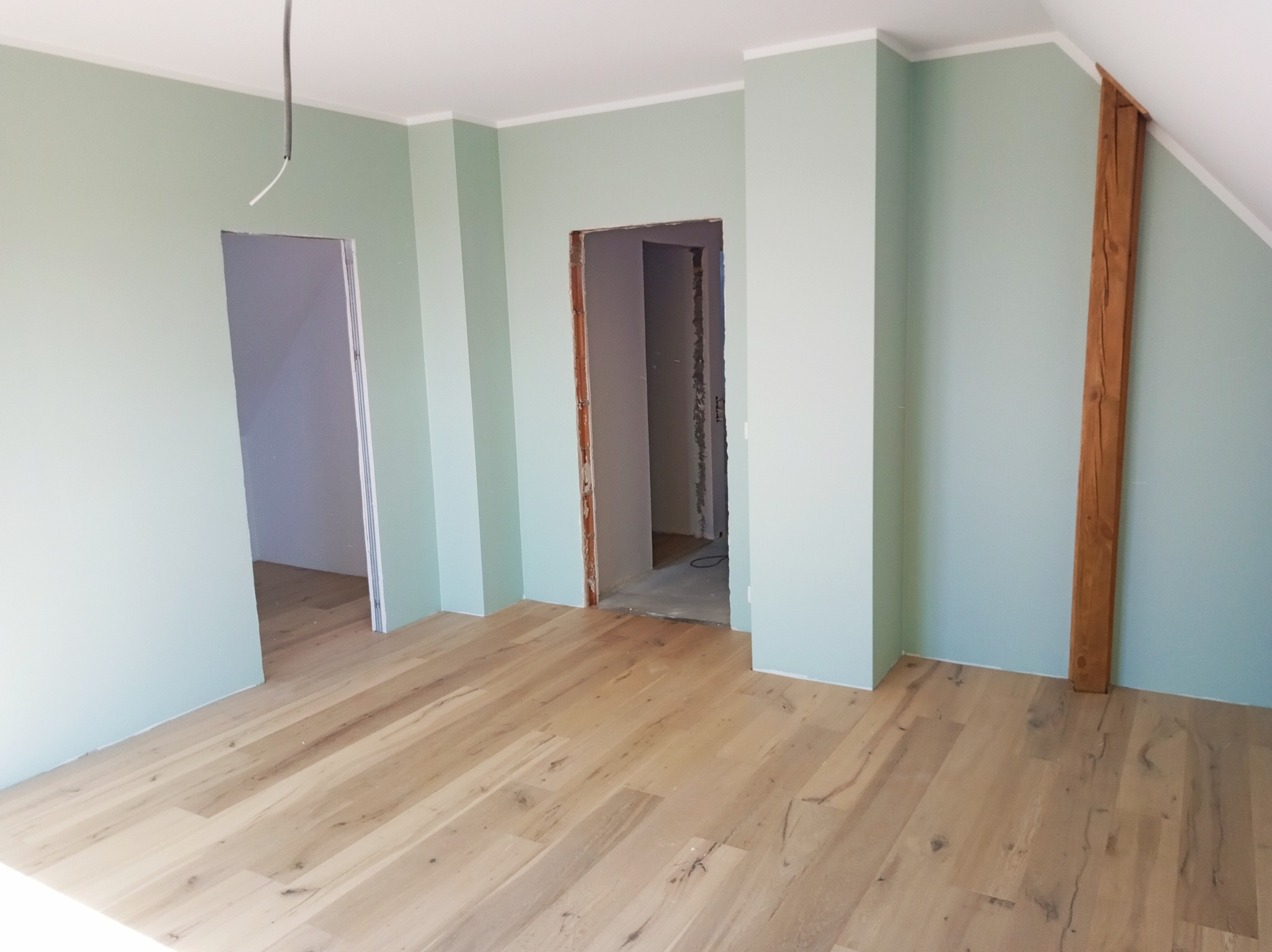 Jasne panele podłogowe w pokoju z pomalowanymi na zielono ścianami i drewnianą belką. Widoczne otwory drzwiowe w trakcie wykończenia.