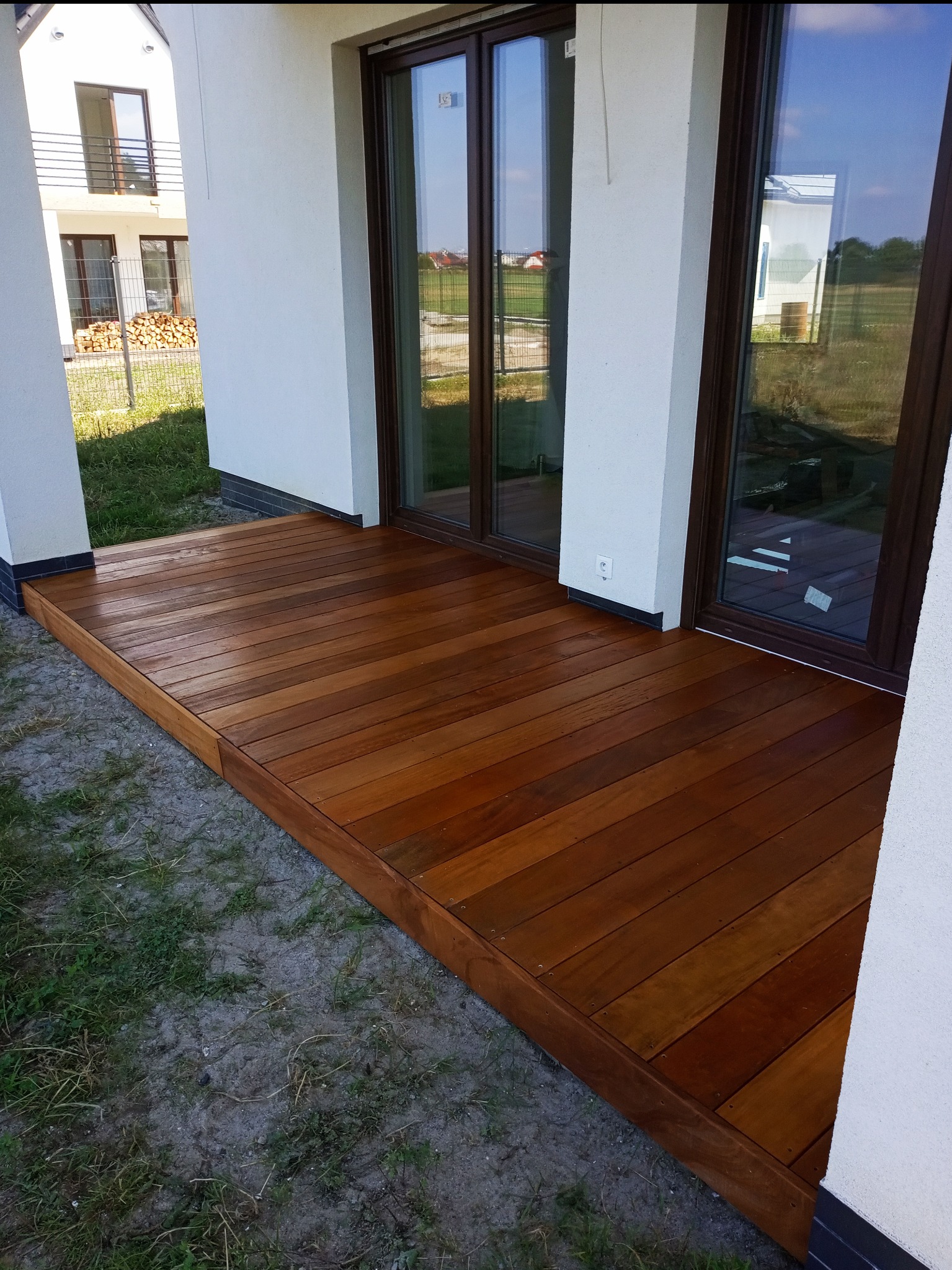 Balkon z drewnianą podłogą, widok z zewnątrz. Brązowe deski ułożone równolegle, białe ściany budynku z brązowymi ramami okiennymi.