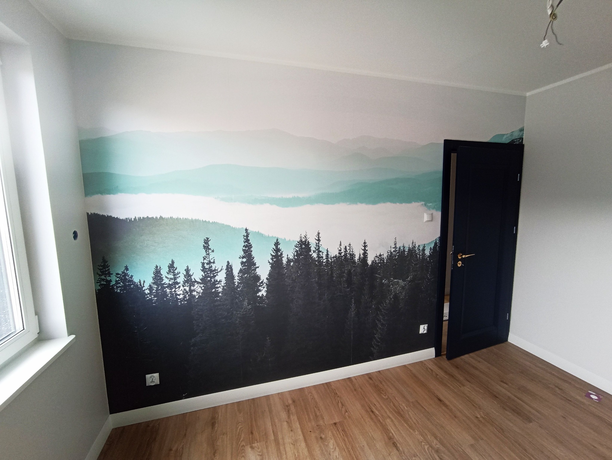 Pokój po remoncie: fototapeta z motywem górskim, drewniana podłoga, ciemne drzwi. Widoczne gniazdka elektryczne na ścianie z tapetą.