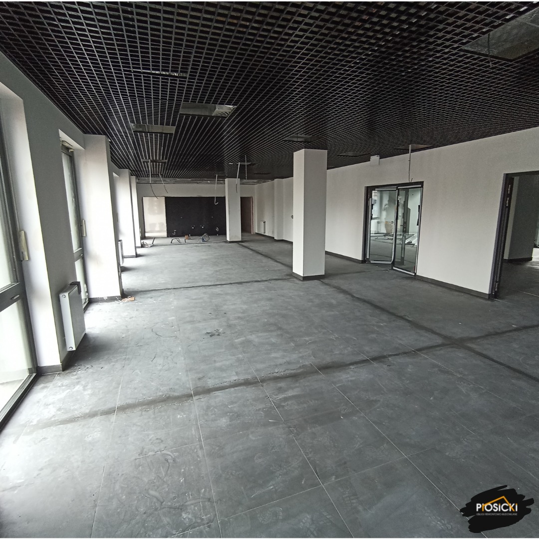 Wnętrze biurowe w trakcie wykończenia: szare płytki na podłodze, białe ściany, czarny sufit podwieszany. Widoczne kolumny i okna. Przestrzeń do adaptacji.