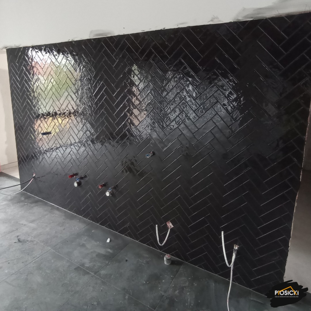 Ściana wyłożona błyszczącymi, czarnymi płytkami ułożonymi we wzór jodełki. Widoczne instalacje wodne i elektryczne, szara podłoga. Częściowo wykończone wnętrze łazienki.