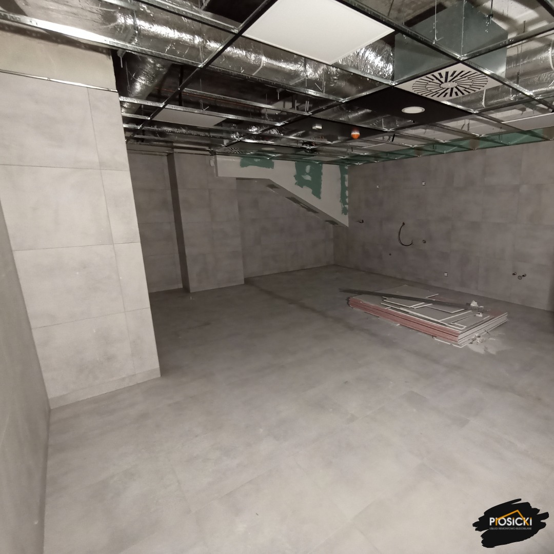Wnętrze w szarościach z ułożoną podłogą z płytek i fragmentem sufitu podwieszanego. Na podłodze leżą płyty gipsowe. Widoczne ślady po instalacjach.