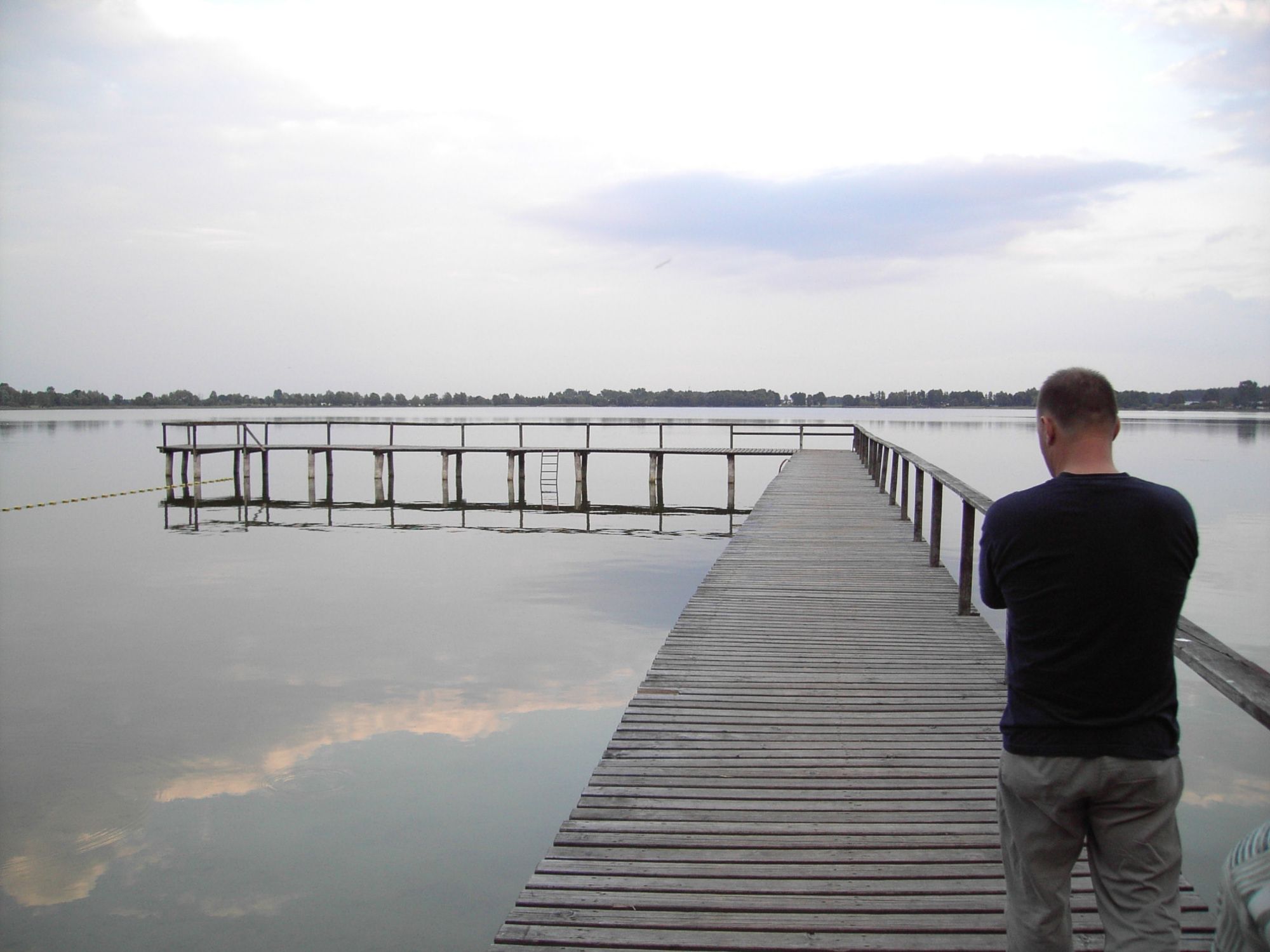Mężczyzna stoi na drewnianym molo nad jeziorem, na wodzie widoczne odbicie chmur, w tle linia drzew
