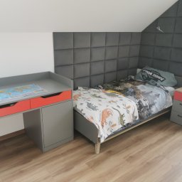 Kupis Kuchnie - Pokój dziecięcy z szarym łóżkiem, tapicerowanym panelem ściennym, biurkiem z czerwonymi szufladami i szafką nocną, całość w jasnej tonacji.