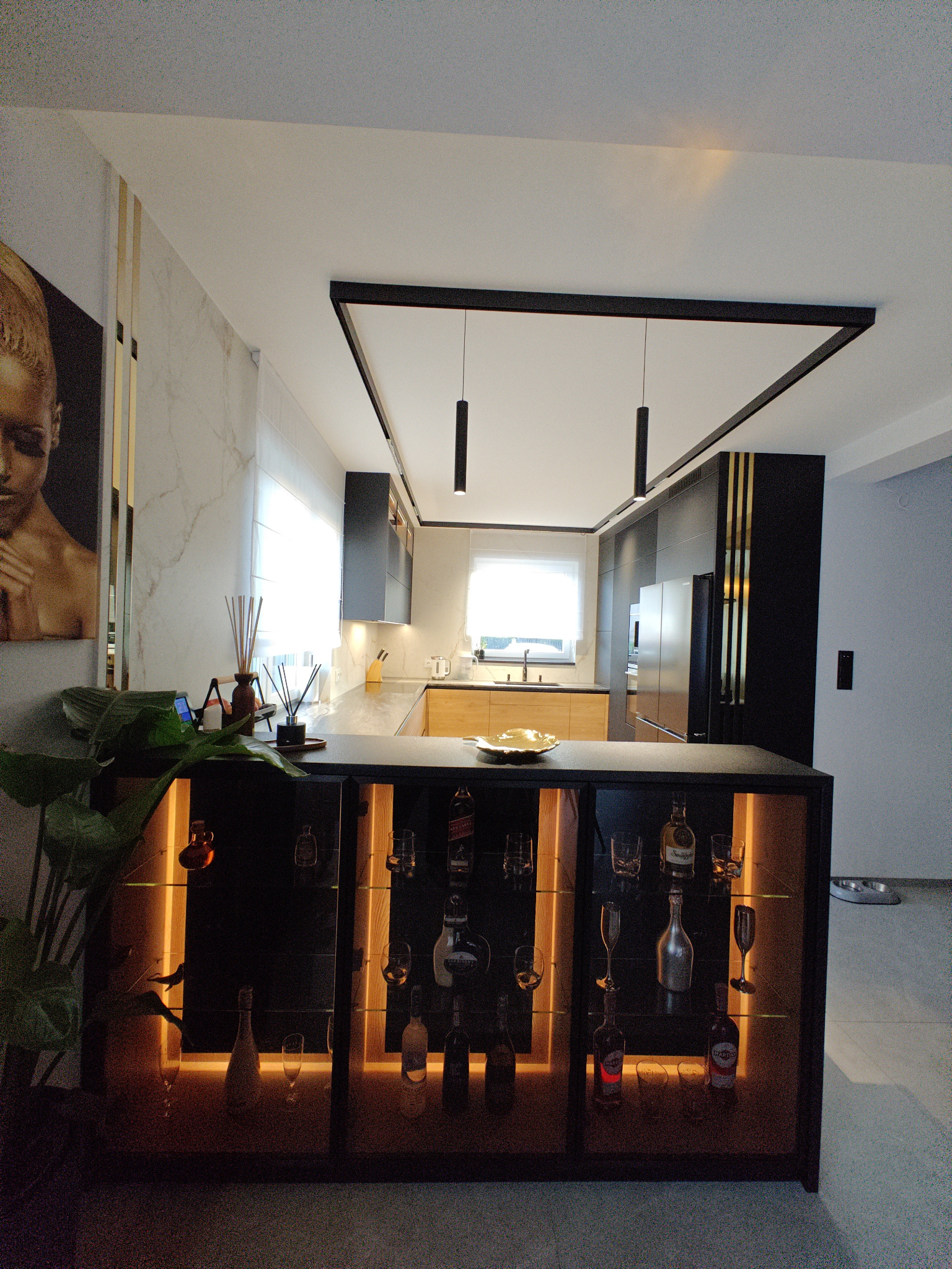 Nowoczesna kuchnia z czarnymi szafkami, drewnianymi blatami, oświetleniem punktowym, wyspą z podświetlaną witryną na alkohole i dekoracyjną grafiką na ścianie.