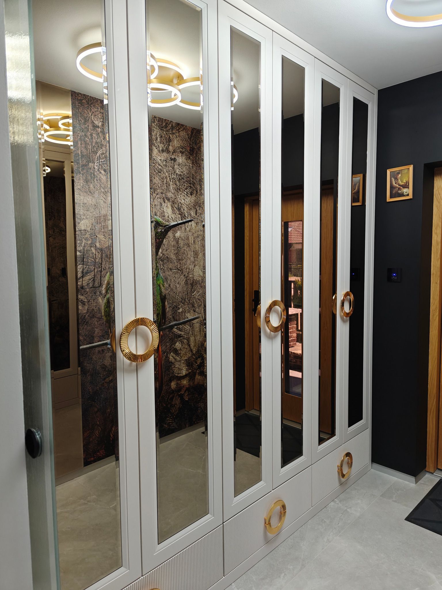 Biała szafa wnękowa z lustrzanymi drzwiami, złotymi okrągłymi uchwytami i dolnymi panelami z pionowymi żłobieniami, odbijająca wnętrze z wzorzystą tapetą i nowoczesnym oświetleniem.