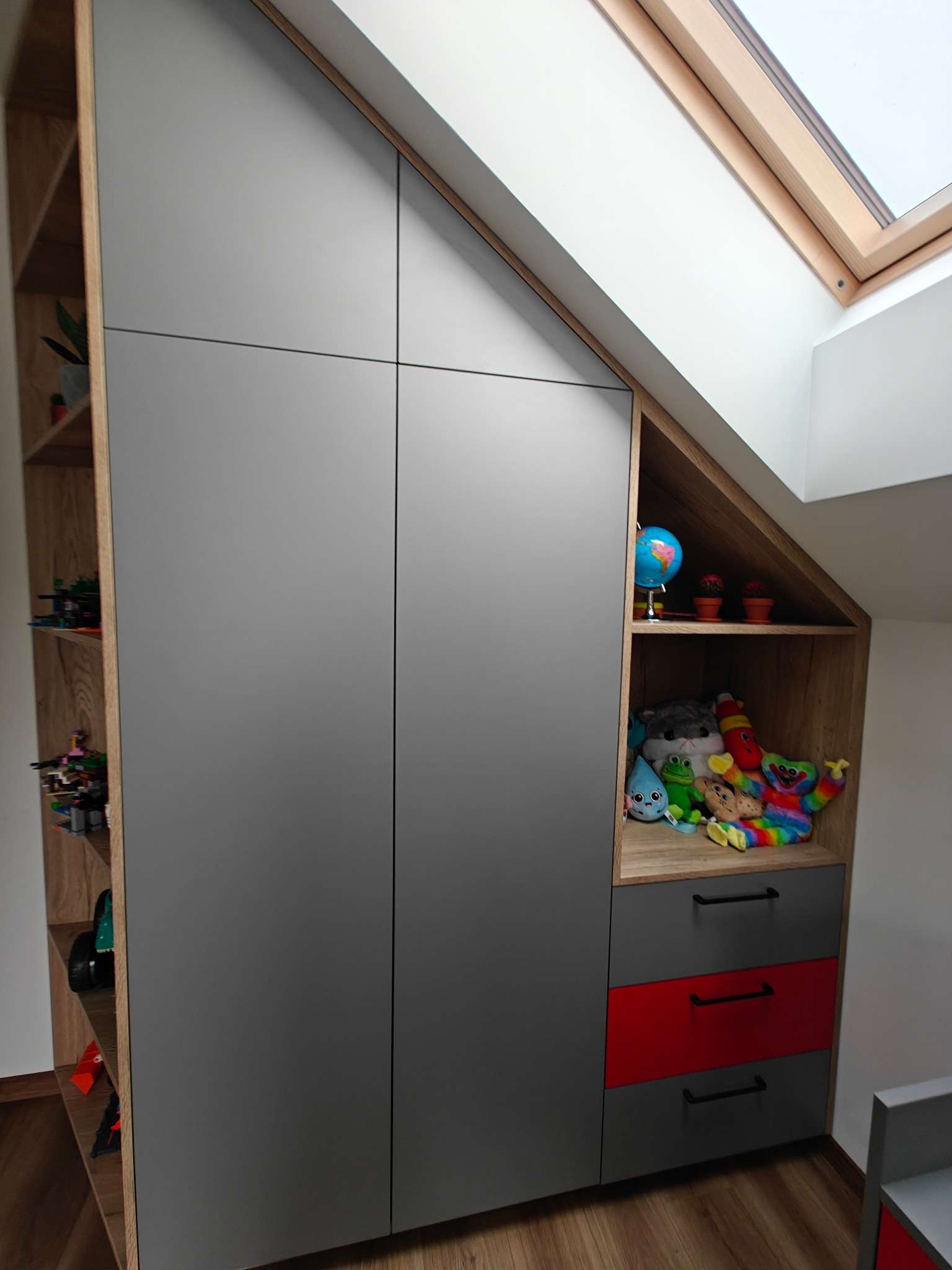 Szafa wnękowa na poddaszu ze skośnym sufitem, szare fronty, drewniane półki z zabawkami, czerwona szuflada.
