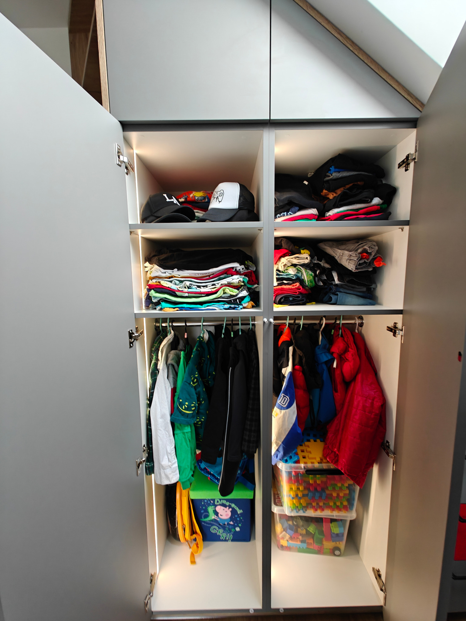 Otwarta szafa wnękowa z szarymi frontami, podświetlona od wewnątrz, wypełniona ubraniami, czapkami i zabawkami w plastikowych pojemnikach.
