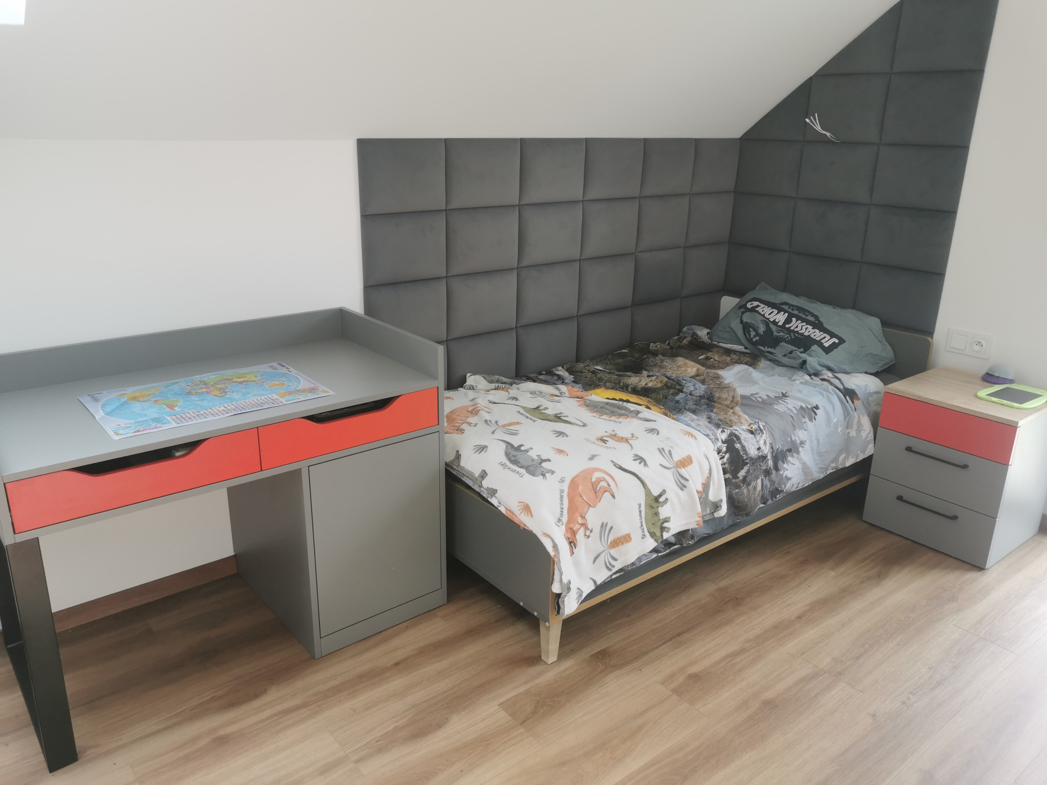 Pokój dziecięcy z szarym łóżkiem, tapicerowanym panelem ściennym, biurkiem z czerwonymi szufladami i szafką nocną, całość w jasnej tonacji.