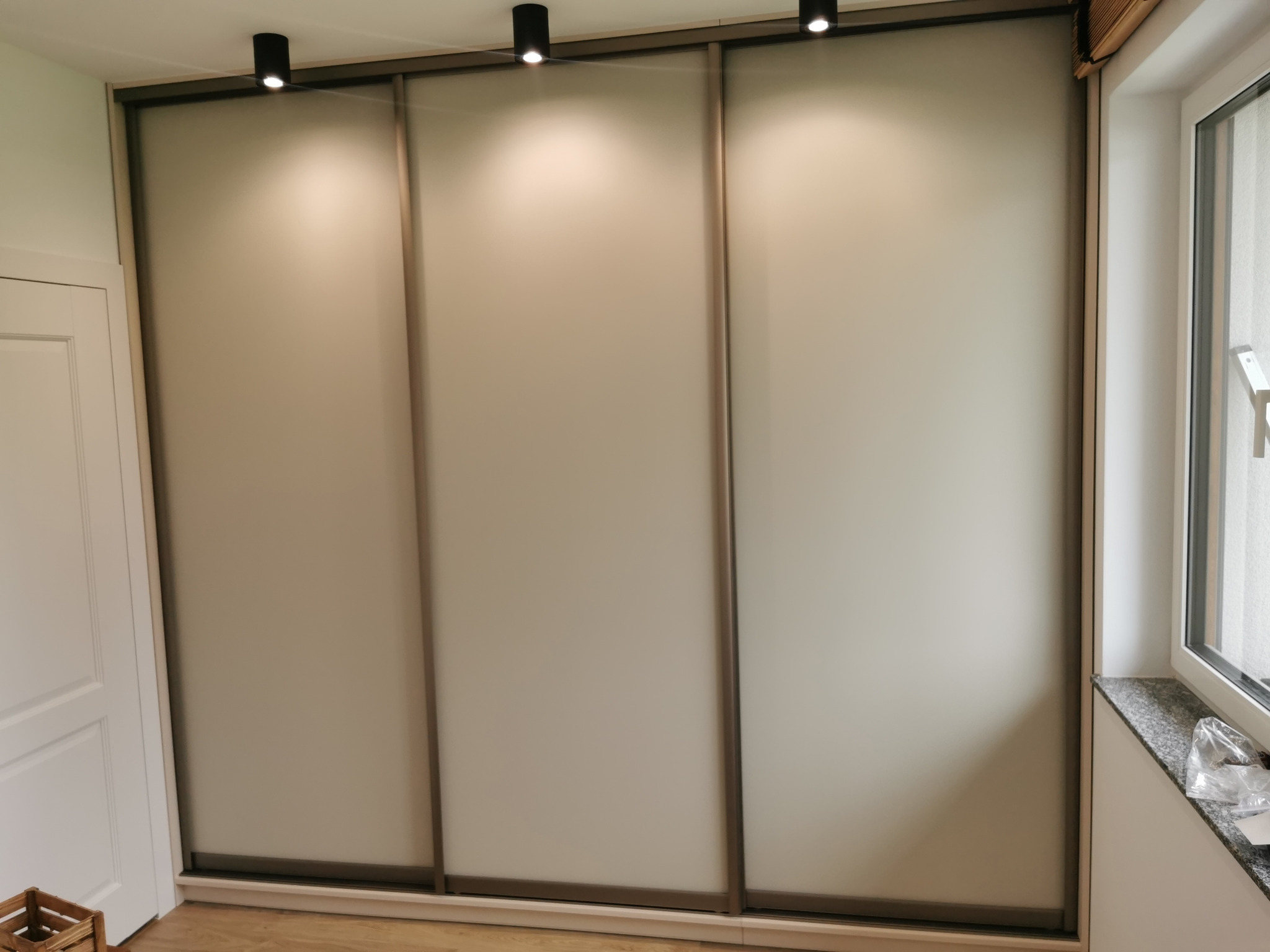 Trzydrzwiowa szafa przesuwna z matowego szkła, oświetlona trzema punktowymi lampami sufitowymi, widoczna obok białych drzwi i okna z marmurowym parapetem.