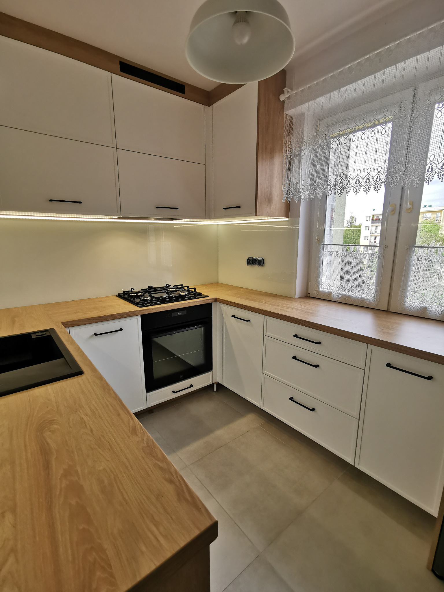 Jasna kuchnia z białymi szafkami i drewnianymi blatami, czarne uchwyty, okno z koronkową firanką, płyta gazowa i piekarnik w rogu, oświetlenie LED pod szafkami wiszącymi.