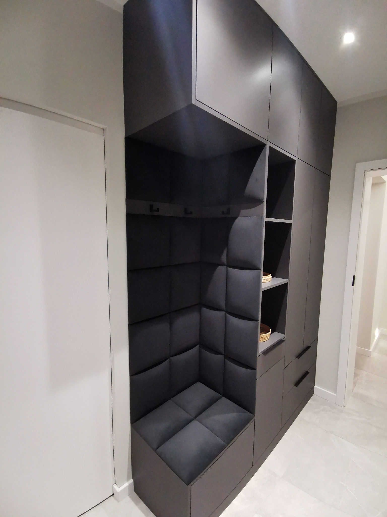 Ciemnoszara szafa wnękowa z siedziskiem tapicerowanym panelami w romby, wbudowana we wnękę w przedpokoju, z widocznymi haczykami na ubrania i półkami z koszykami.