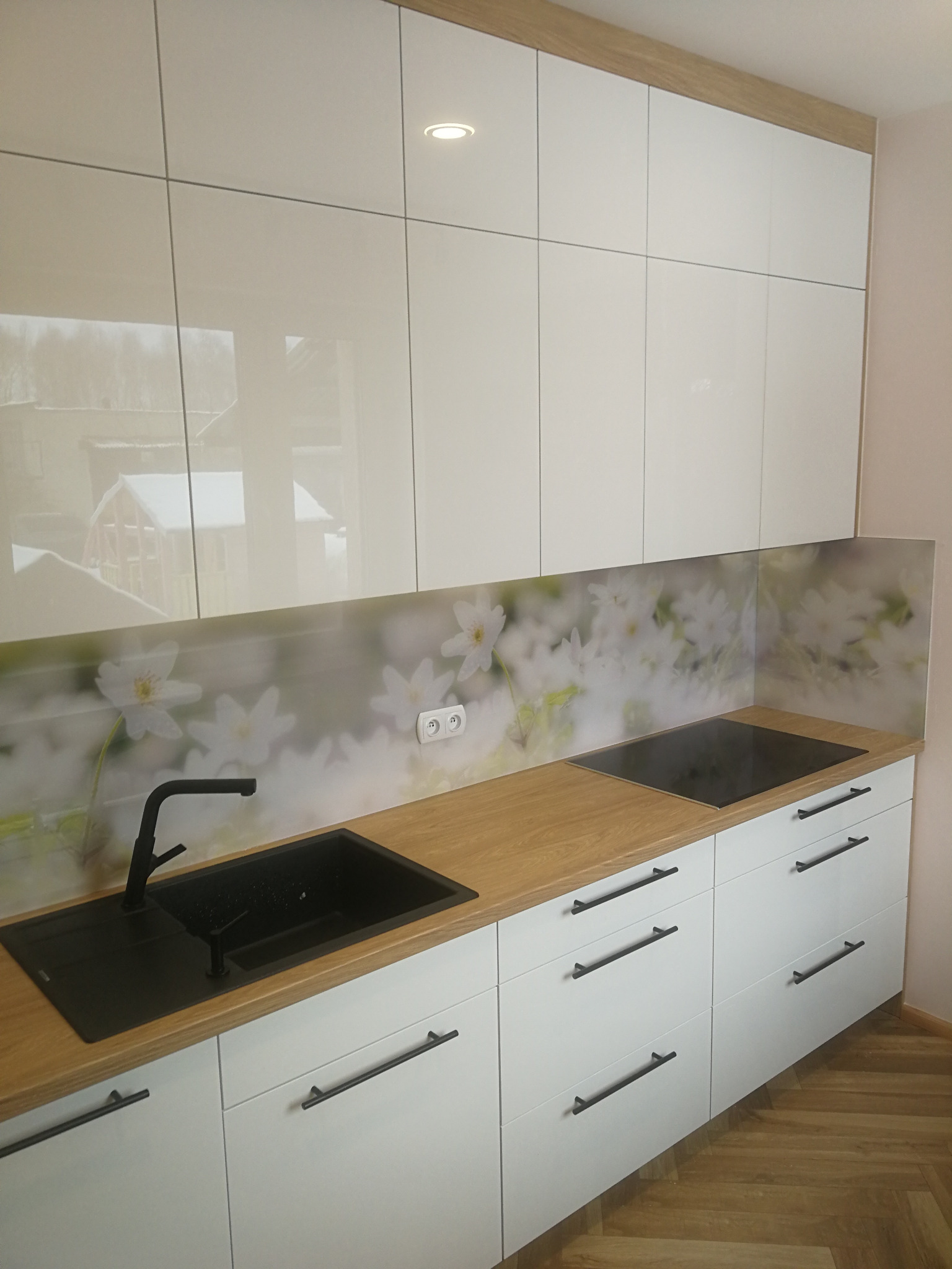 Biała kuchnia z drewnianym blatem i nadrukiem kwiatowym na szkle, czarny zlew i bateria, minimalistyczne uchwyty, parkiet w jodełkę.