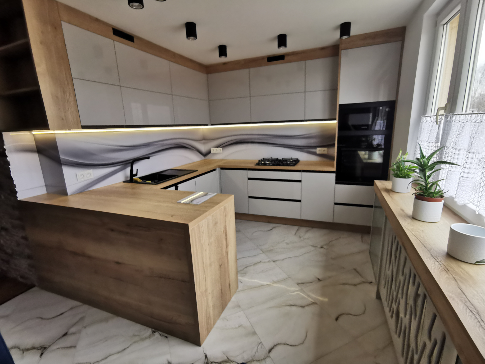 Nowoczesna kuchnia z wyspą, białe fronty szafek z drewnianymi akcentami, czarne oświetlenie punktowe, marmurowe płytki na podłodze, rośliny doniczkowe na parapecie.