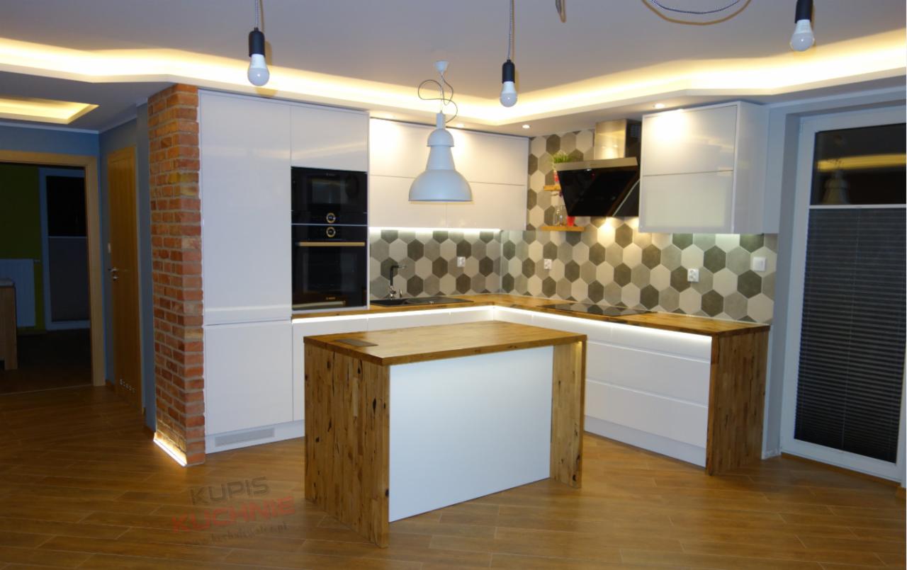Nowoczesna kuchnia z białymi lakierowanymi szafkami, drewnianym blatem i wyspą, oświetleniem LED pod szafkami i lampami wiszącymi, ściana z dekoracyjnej cegły i heksagonalne płytki na ścianie.