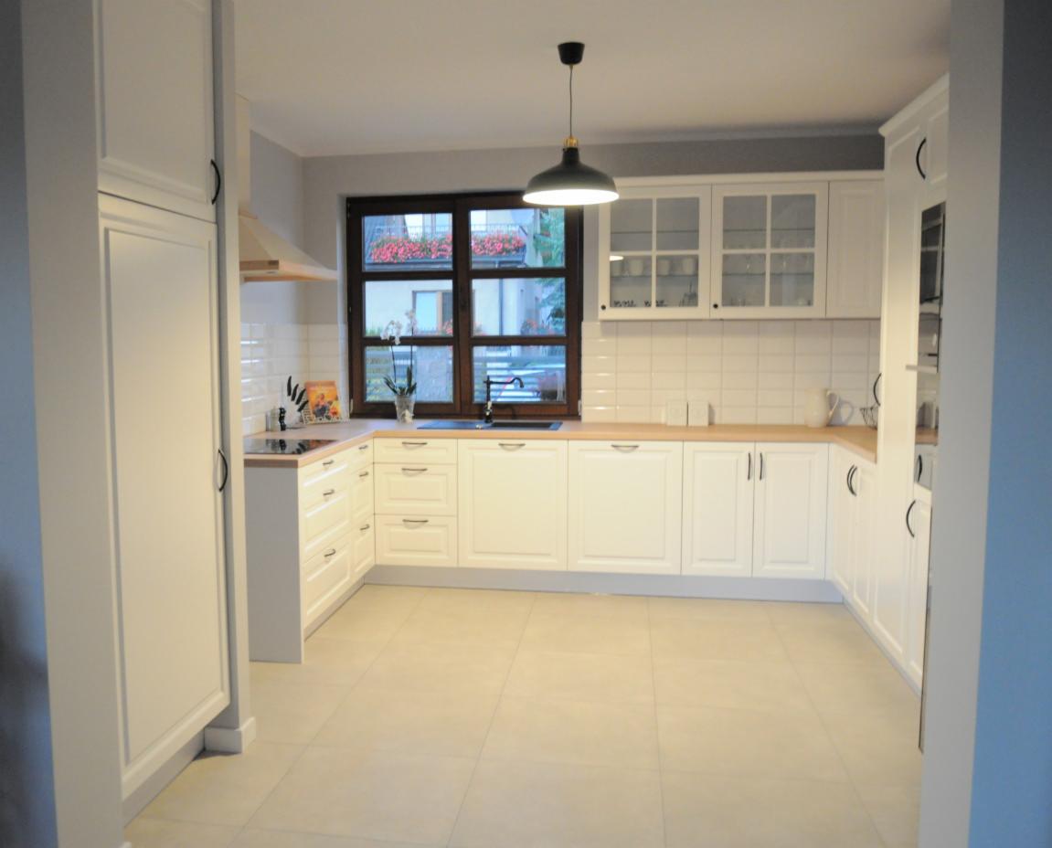 Jasna, przestronna kuchnia z białymi szafkami, drewnianym blatem i dużym oknem z widokiem na zieleń oraz kwiaty w donicach. Nad blatem wiszą szafki z przeszklonymi frontami. Podłoga wyłożona jest...