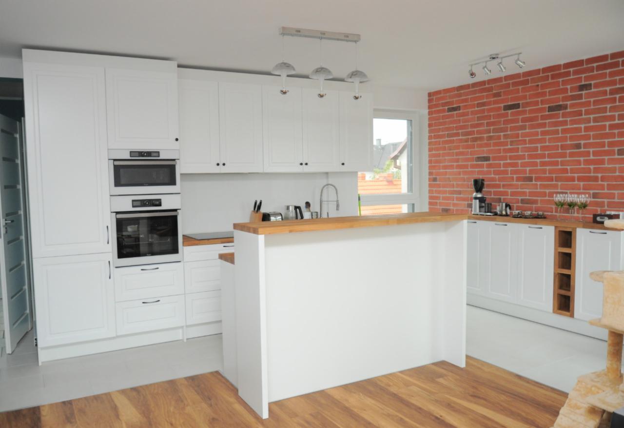Jasna kuchnia z białymi frontami szafek, drewnianym blatem, wyspą kuchenną i ścianą z czerwonej cegły. Widoczny sprzęt AGD w zabudowie i drewniana podłoga.