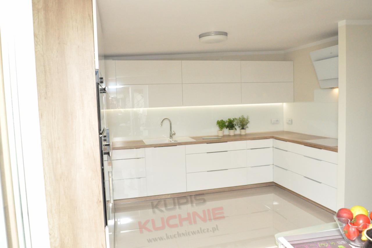 Nowoczesna, jasna kuchnia z białymi, błyszczącymi frontami szafek, drewnianym blatem i oświetleniem LED pod górnymi szafkami. Na blacie stoją małe doniczki z zielonymi roślinami. Na podłodze...