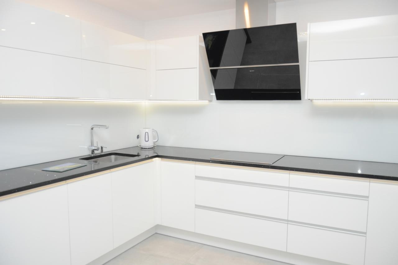 Nowoczesna, biała kuchnia z czarnym blatem i minimalistycznym okapem, oświetleniem LED pod szafkami i czajnikiem elektrycznym na blacie.