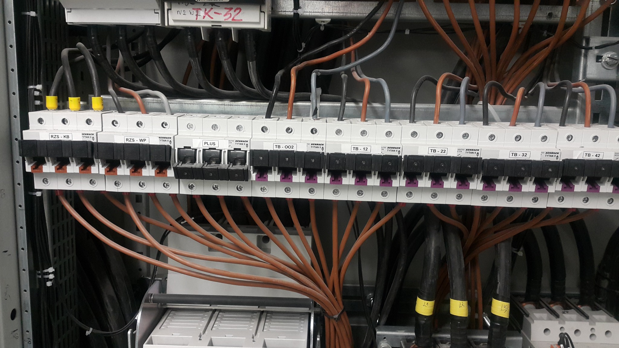 Rozdzielnica elektryczna z widocznymi bezpiecznikami, przewodami o różnych kolorach (brązowy, czarny, szary) i oznaczeniami na listwach zaciskowych, wewnątrz metalowej obudowy.