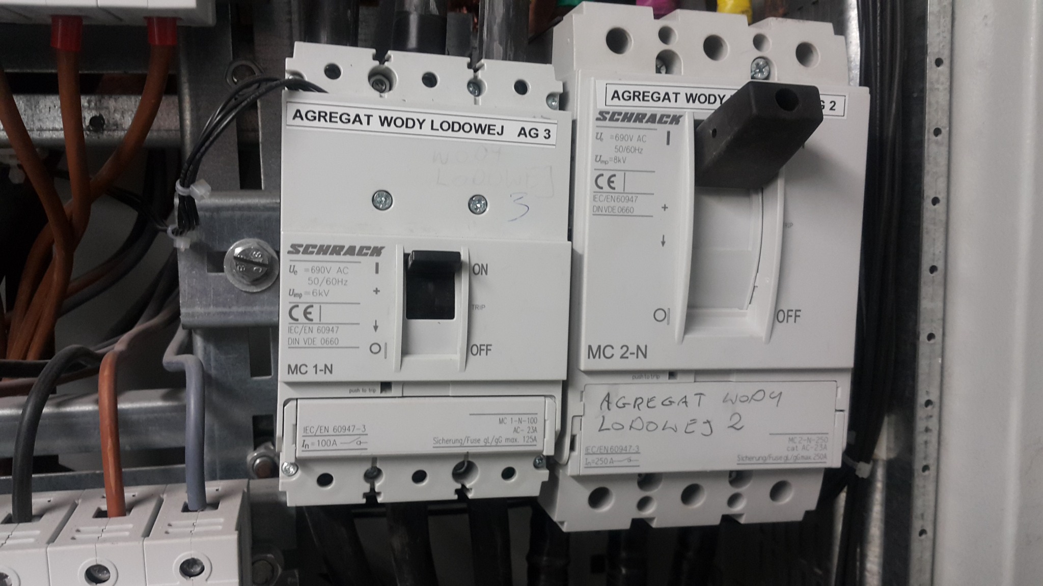 Rozdzielnica elektryczna z wyłącznikami marki Schrack, opisane jako 'Agregat wody lodowej', z widocznymi przewodami i oznaczeniami technicznymi, zamontowana w metalowej obudowie.