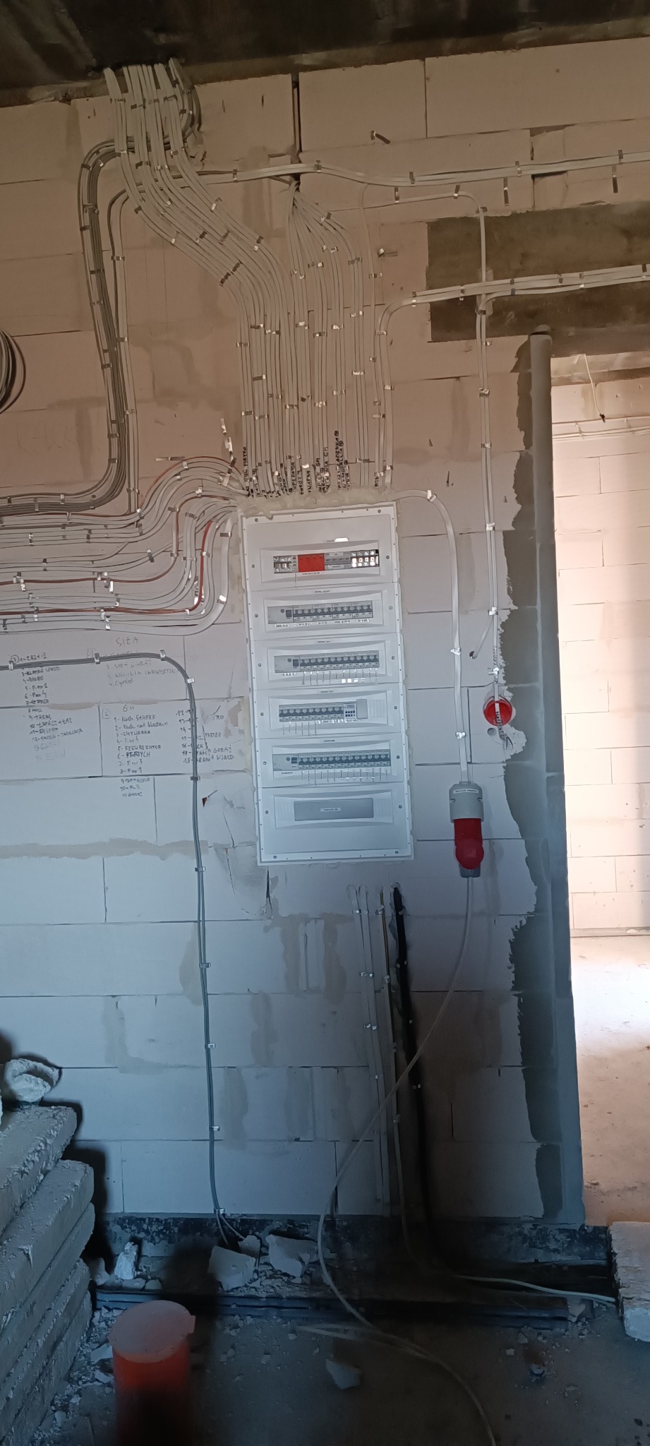 Rozdzielnia elektryczna z licznymi przewodami w peszlach, instalacja w surowym stanie, ściana z bloczków, widoczne notatki na ścianie obok rozdzielni, gniazdo siłowe w kolorze czerwonym