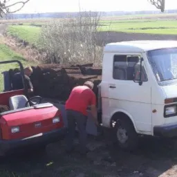Pracownicy rozładowują z białego samochodu dostawczego rolki trawy na przyczepkę wózka transportowego na tle zielonego pola.