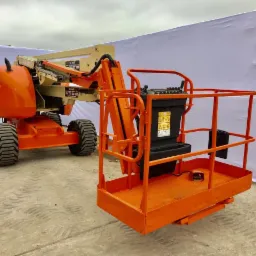 Pomarańczowy podnośnik koszowy z wysięgnikiem na nierównym terenie, z widocznymi oznaczeniami bezpieczeństwa, na tle białej ściany.