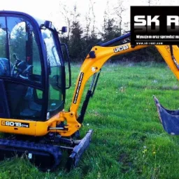Żółta minikoparka JCB 8018CTS na gąsienicach, stojąca na trawiastym terenie, z widocznym logo firmy SK Rent w tle.