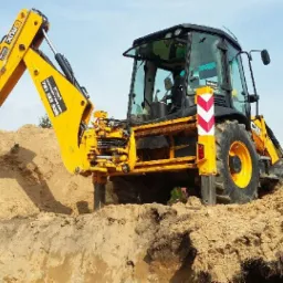 Żółta koparko-ładowarka JCB zagłębia się w ziemię, tworząc wykop, z widocznym numerem telefonu na ramieniu koparki i odblaskowym słupkiem bezpieczeństwa obok koła.