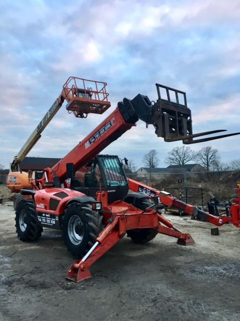 Pomarańczowa ładowarka teleskopowa Manitou z wysięgnikiem i platformą roboczą, stojąca na placu budowy pod pochmurnym niebem.