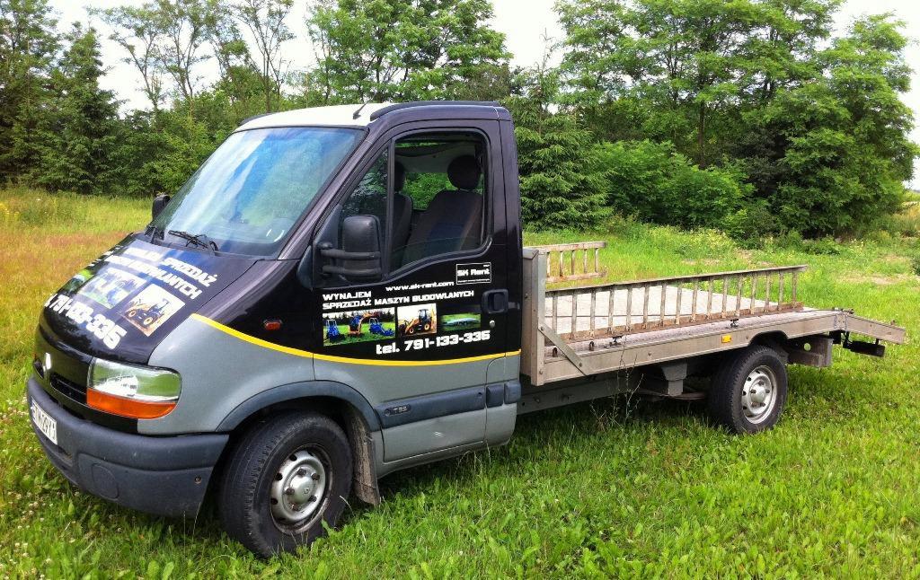 Czarno-szary samochód dostawczy z platformą transportową, oklejony reklamami maszyn budowlanych, zaparkowany na trawiastym terenie.