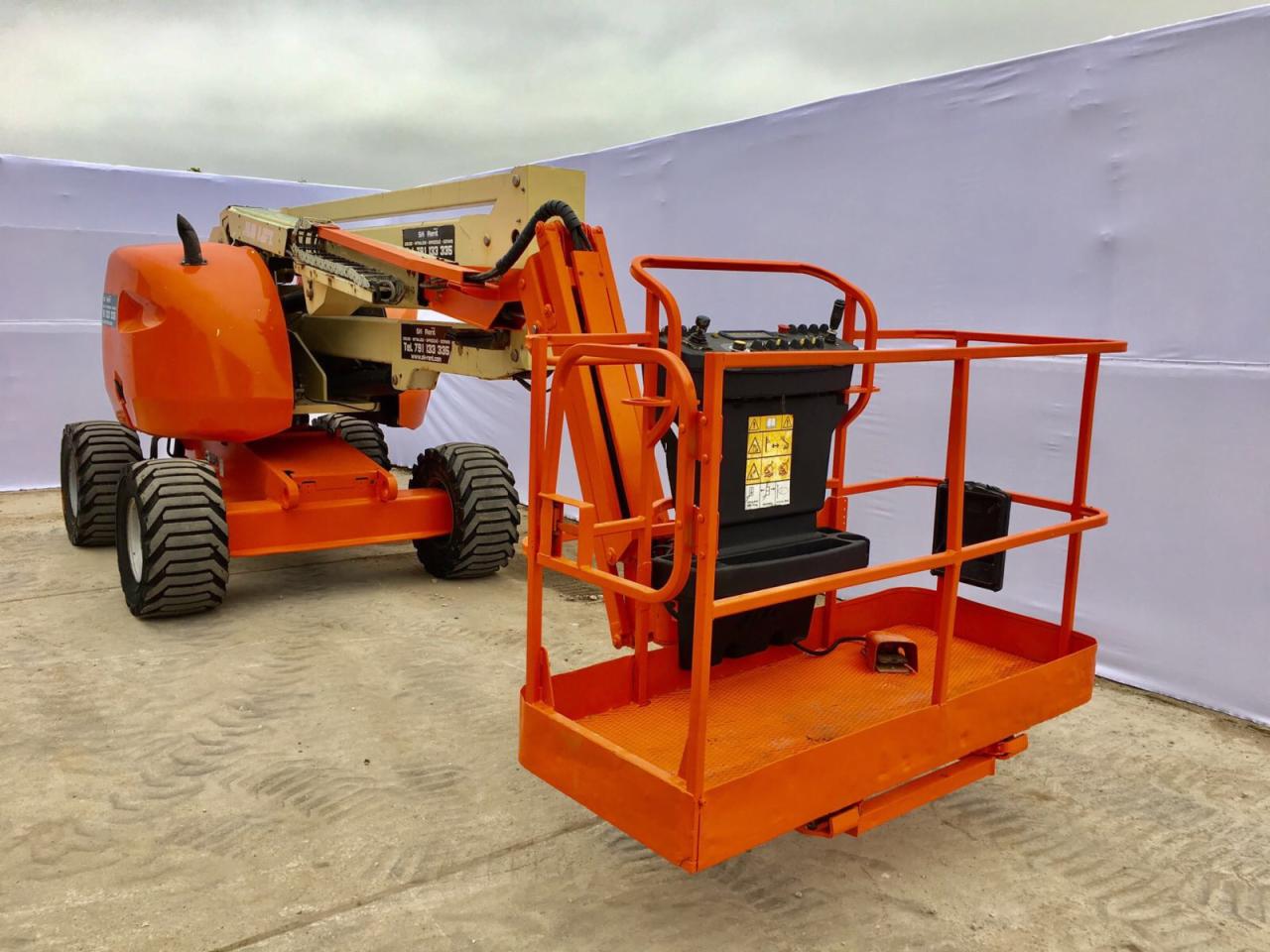 Pomarańczowy podnośnik koszowy z wysięgnikiem na nierównym terenie, z widocznymi oznaczeniami bezpieczeństwa, na tle białej ściany.