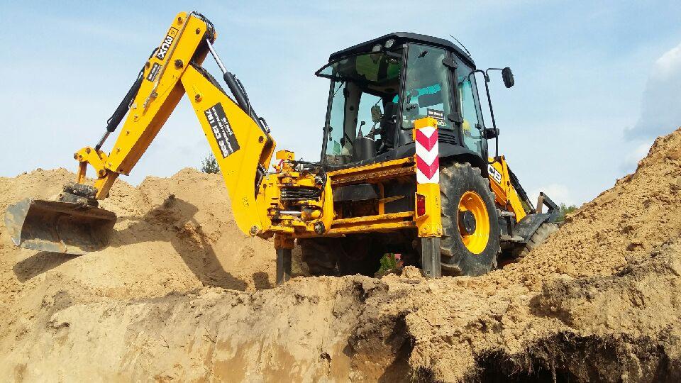Żółta koparko-ładowarka JCB zagłębia się w ziemię, tworząc wykop, z widocznym numerem telefonu na ramieniu koparki i odblaskowym słupkiem bezpieczeństwa obok koła.
