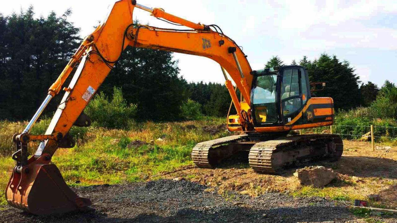 Pomarańczowa koparka JCB na gąsienicach, stojąca na placu budowy z widocznymi śladami użytkowania, z lasem w tle.