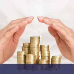 Dłonie kobiety w białej koszuli osłaniają stosy złotych monet na ciemnoniebieskim tle, symbolizujące bezpieczeństwo finansowe.