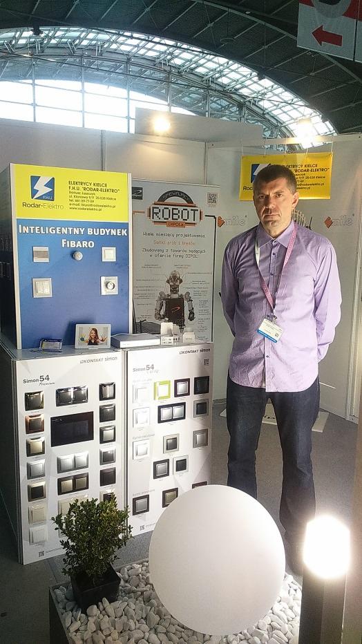 Stoisko z ekspozycją inteligentnych systemów Fibaro i osprzętu Simon 54 na targach. Mężczyzna w koszuli, prezentacja oferty Rodar-Elektro.