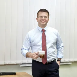 Uśmiechnięty mężczyzna w okularach, błękitnej koszuli i czerwonym krawacie, trzymający kubek z kawą w biurze z żaluzjami w tle.